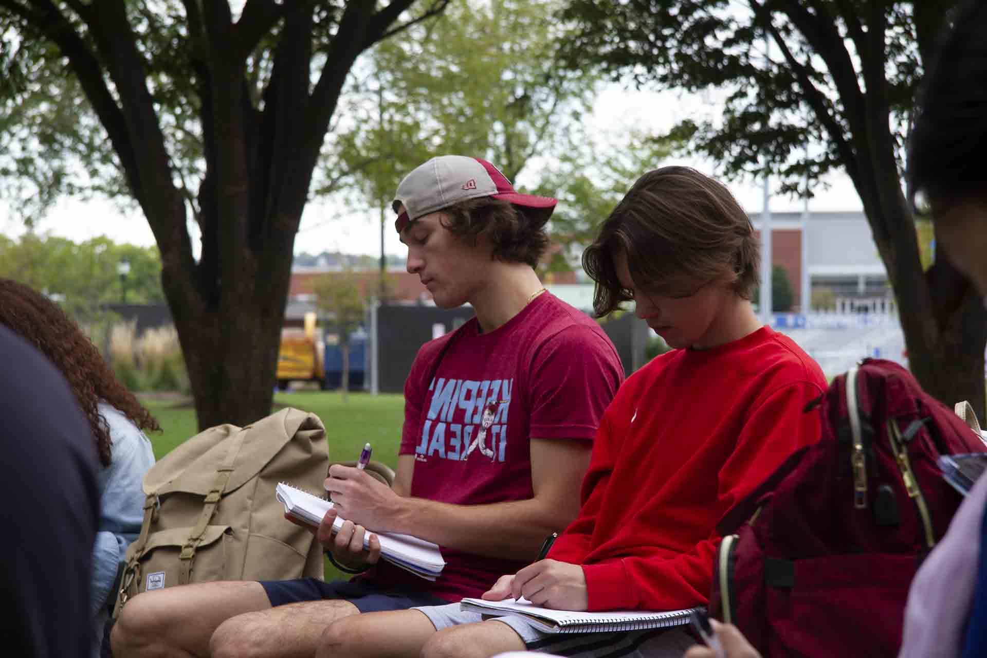 Students take notes during a class session under a tree.