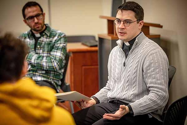 A young Jesuit wearing a priest’s collar leads a circle discussion in a classroom.