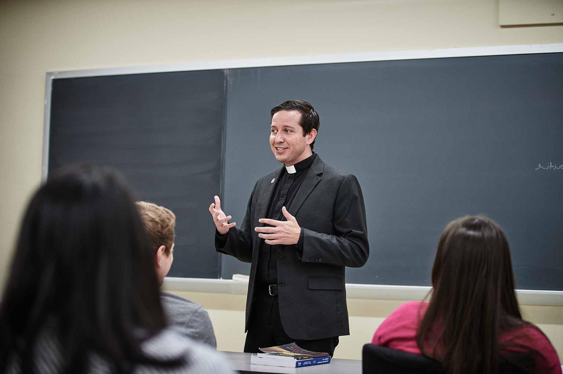 A Jesuit faculty member speaks to students in front of a classroom with a blackboard. The heads of students can be seen in the foreground.