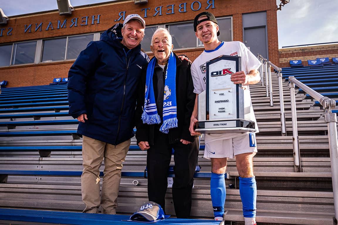 An elderly man, a middle aged man and a young man — the Kleins — pose for a photo with a men's A-10 Championship trophy in the stands of Hermann Stadium.